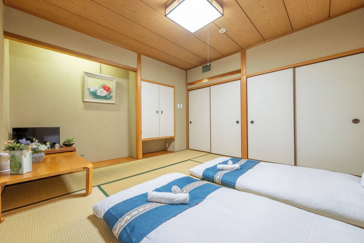 Une pièce de style japonais traditionnel, avec un sol recouvert de tatamis, deux lits avec une literie bleue et blanche, un bureau en bois avec une télévision, et une œuvre d’art florale encadrée accrochée au mur.