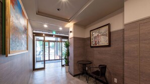 A modern hotel lobby with a large glass door entrance, a potted plant, a small round table with chairs, and a framed picture on the wall.