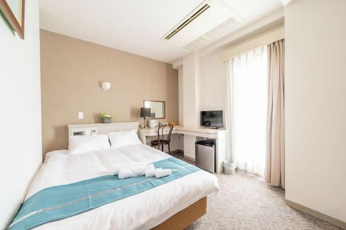 A hotel room with a bed, desk, chair, television, and a window with curtains.