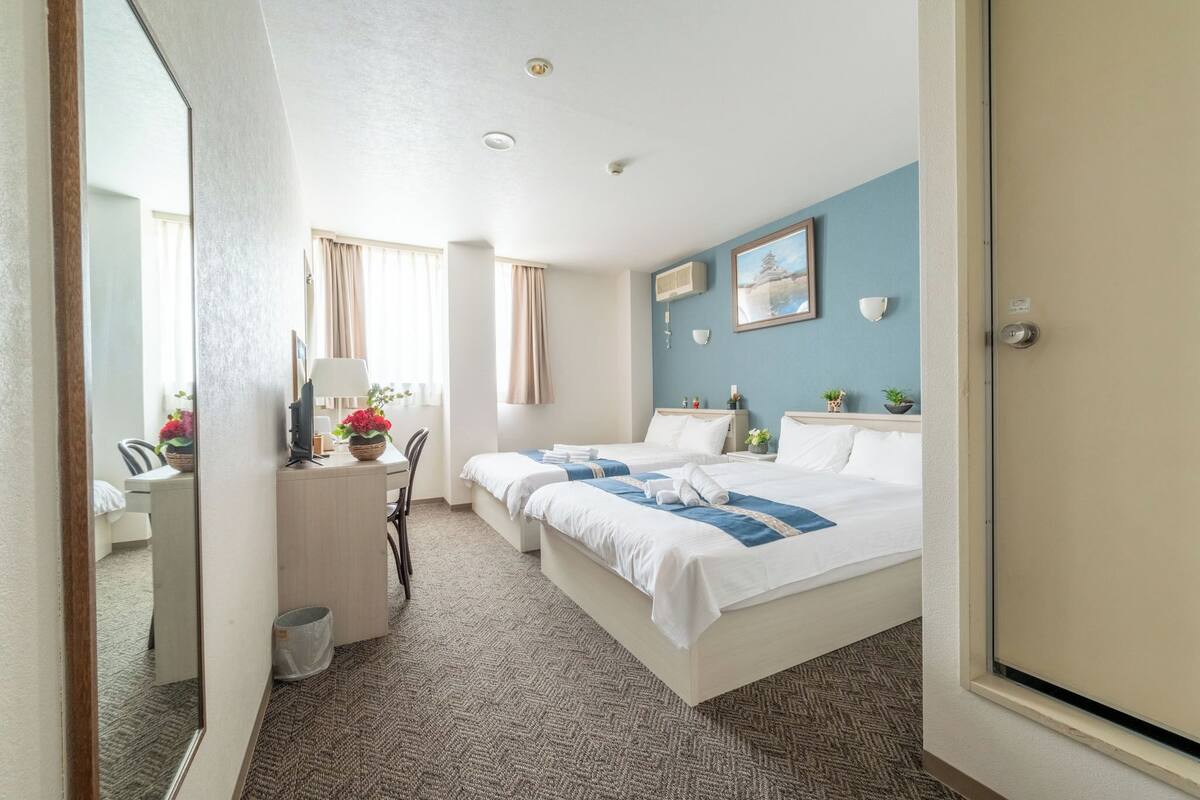 Une chambre d’hôtel avec deux lits, un bureau avec une chaise, une petite table avec une lampe et une fenêtre avec des rideaux.