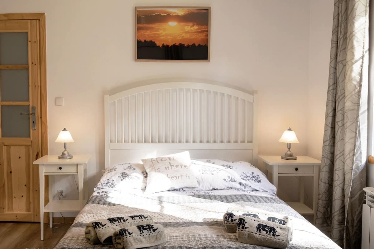 Un dormitorio con puerta de madera, una cama con cabecera blanca, dos mesitas de noche con lámparas y un cuadro de un atardecer en la pared.