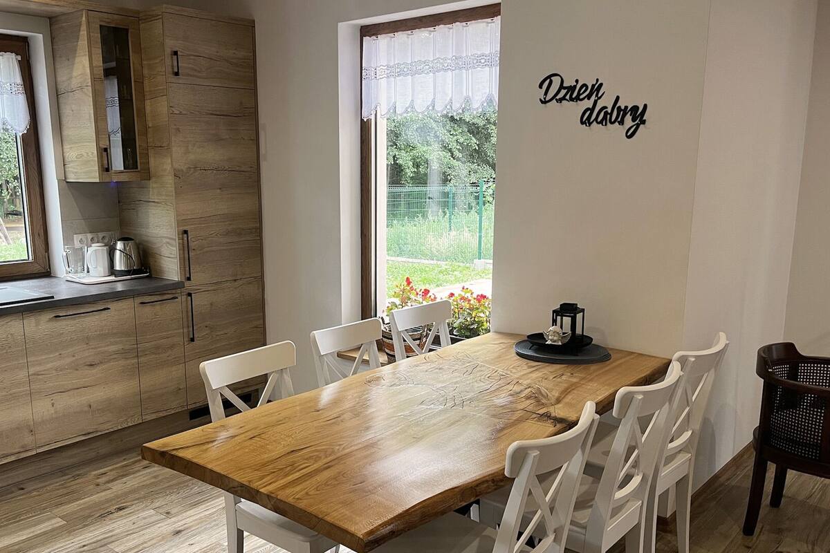 Una cocina moderna con armarios de madera, una mesa de comedor de madera y sillas blancas. Se ve una ventana con vistas a zonas verdes.