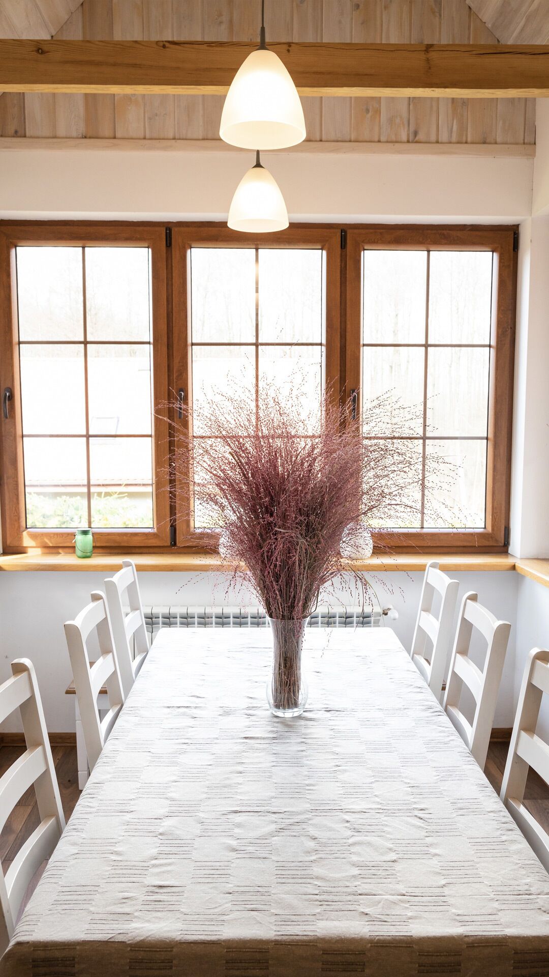 A dining area with a long table, white chairs, and a vase with dried plants. There are wooden beams on the ceiling and large windows with wooden frames.