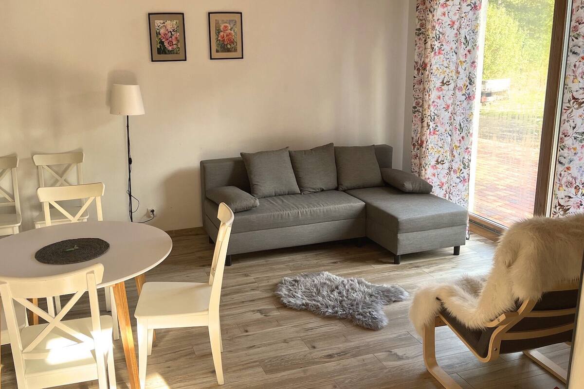Una sala de estar con un sofá gris, sillas blancas, una mesa redonda, una lámpara y cortinas con motivos florales.