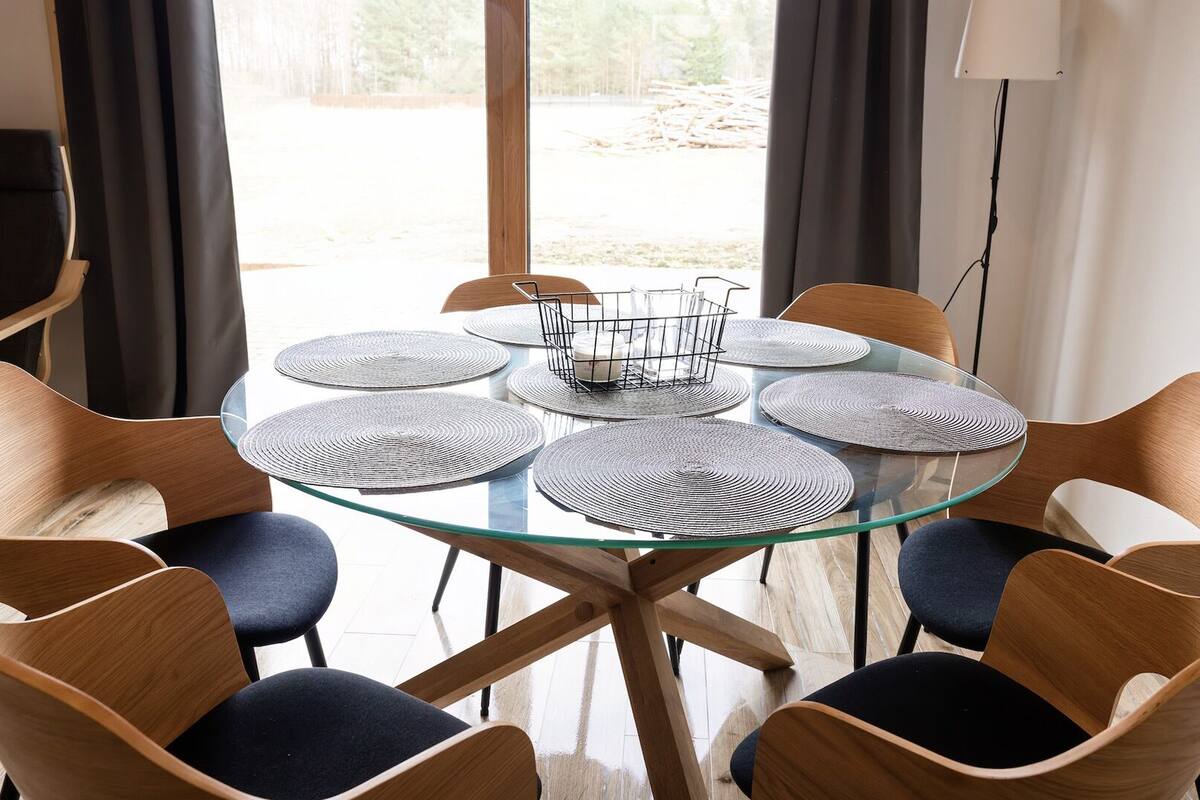 Una mesa redonda de vidrio con seis sillas, cada una con un mantel tejido, en una habitación con vista a una obra en construcción.