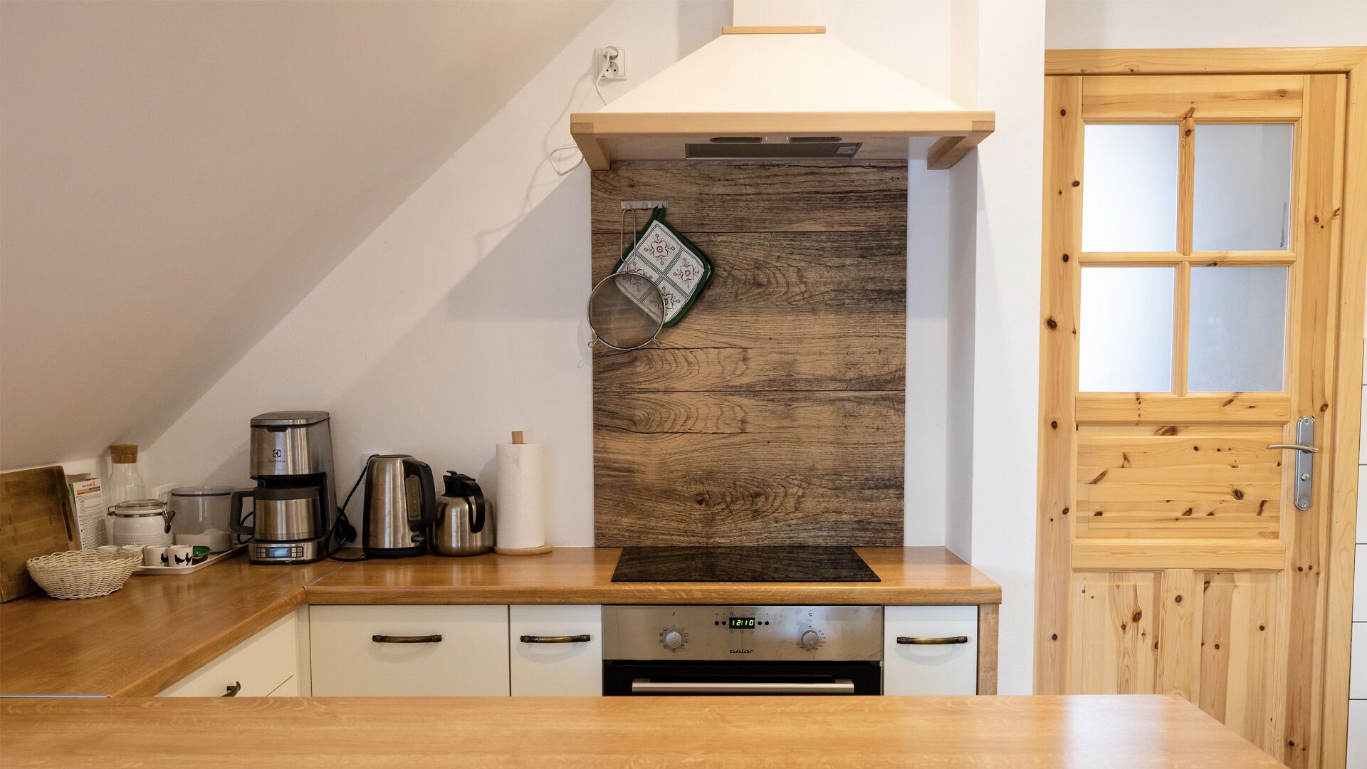 Une cuisine avec des plans de travail en bois, une cuisinière, une machine à café et une horloge indiquant 12h10.
