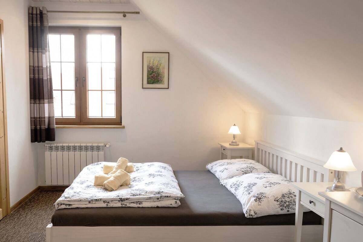 Un dormitorio con cama, dos mesitas de noche, una ventana con cortinas, un radiador y un cuadro enmarcado en la pared.