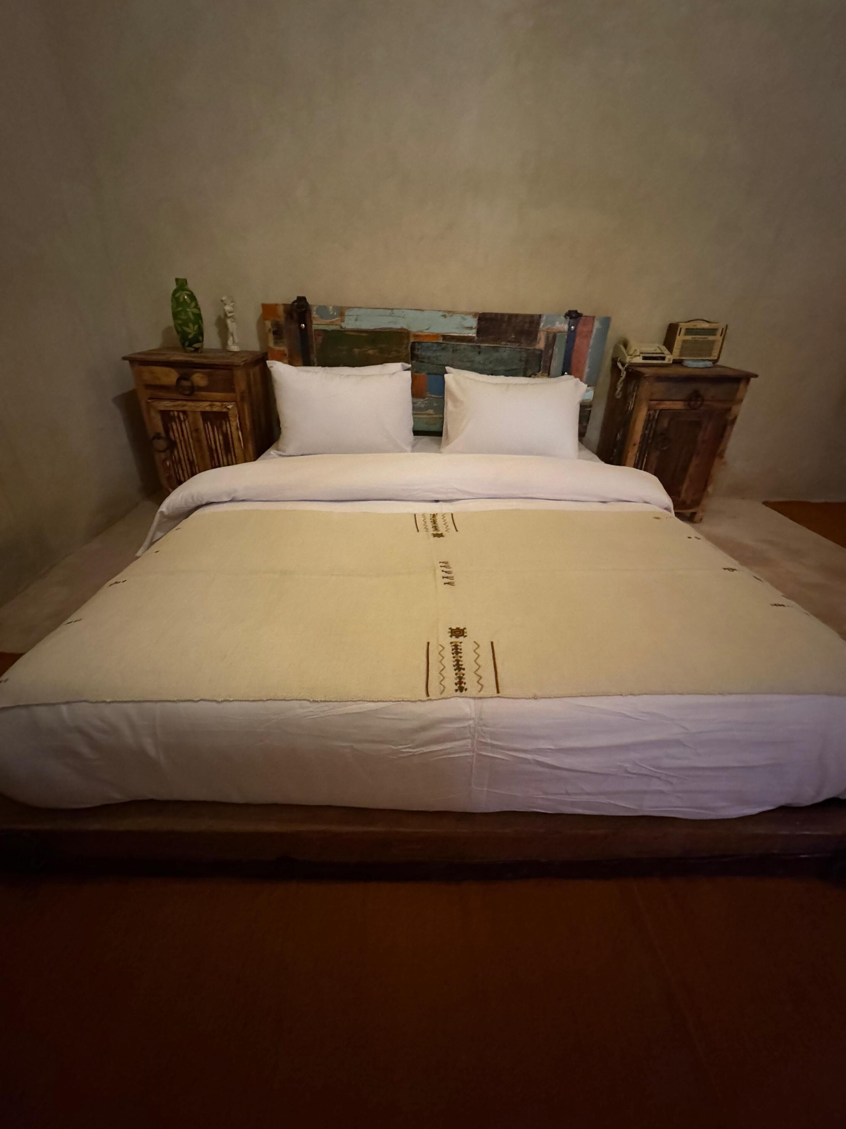 A neatly made bed with white linens, two wooden nightstands with a radio and a green bottle on one, and a brick-patterned headboard.