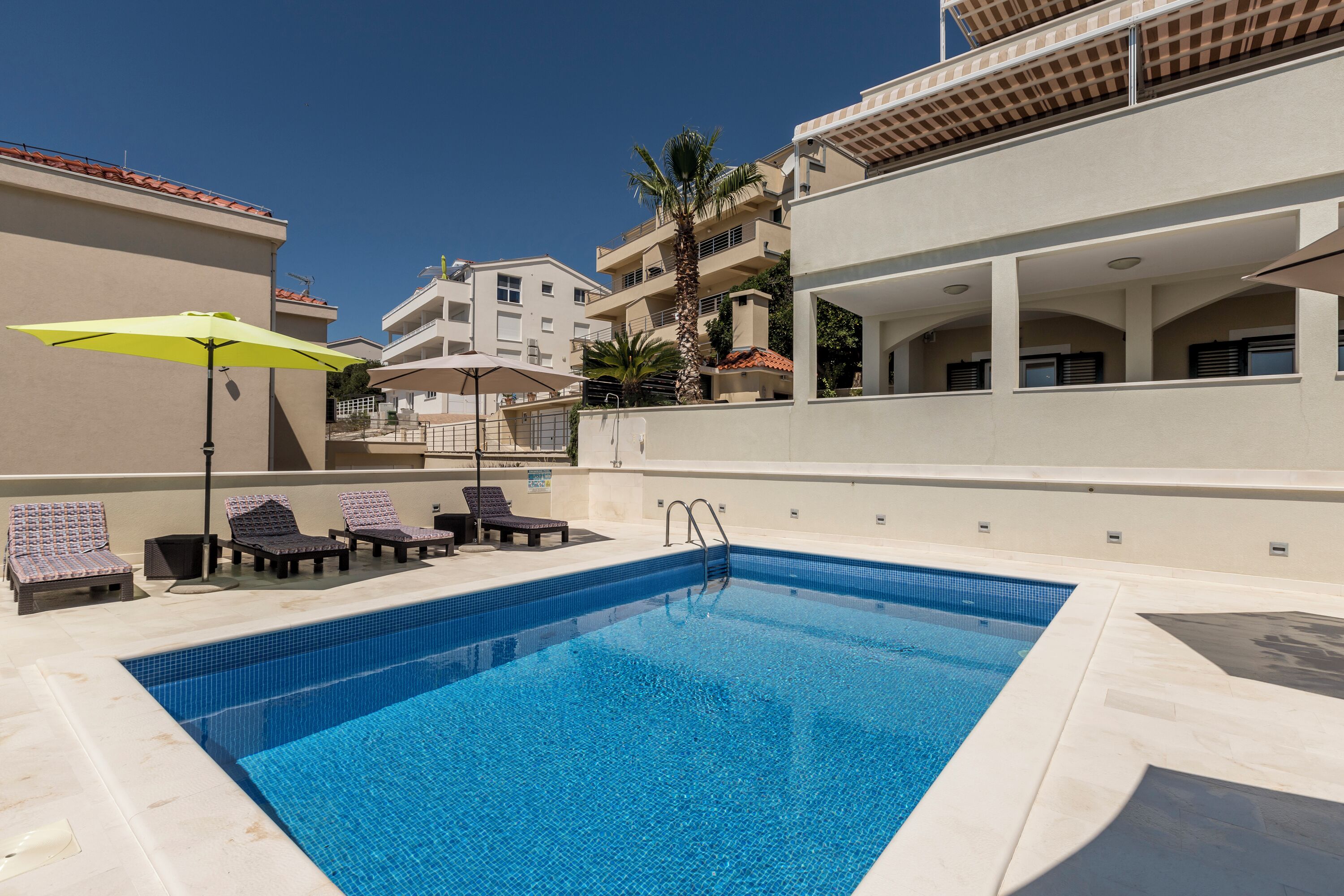 Una piscina con sillones de descanso y sombrillas, rodeada de edificios con balcones y un cielo despejado.