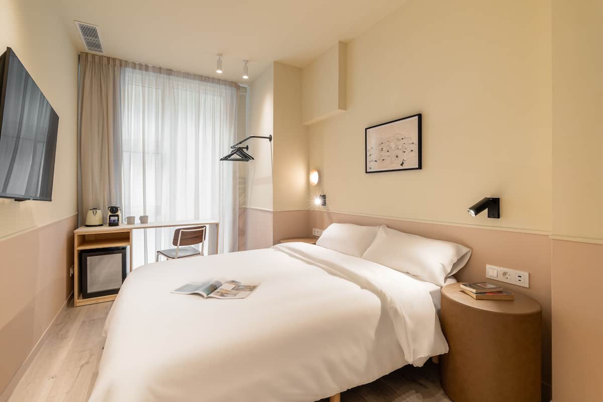 Een hotelkamer met een bed, nachtkastje, lamp en een klein bureau met stoel.
