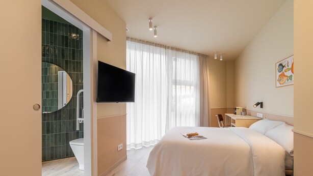 A bedroom with a large bed, a TV mounted on the wall, a bathroom with a toilet and shower, and a window with sheer curtains.