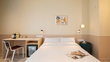 Habitación de hotel con cama, escritorio, silla, refrigerador pequeño, mesita de noche con lámpara y una obra de arte en la pared.