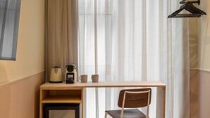 A hotel room with a wooden desk, a chair, a bed, a small table with a kettle, cups, and a book.