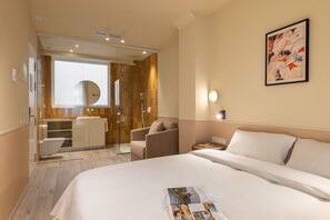 A hotel room with a large bed, a desk, a chair, a glass-enclosed shower, and a wall-mounted artwork.