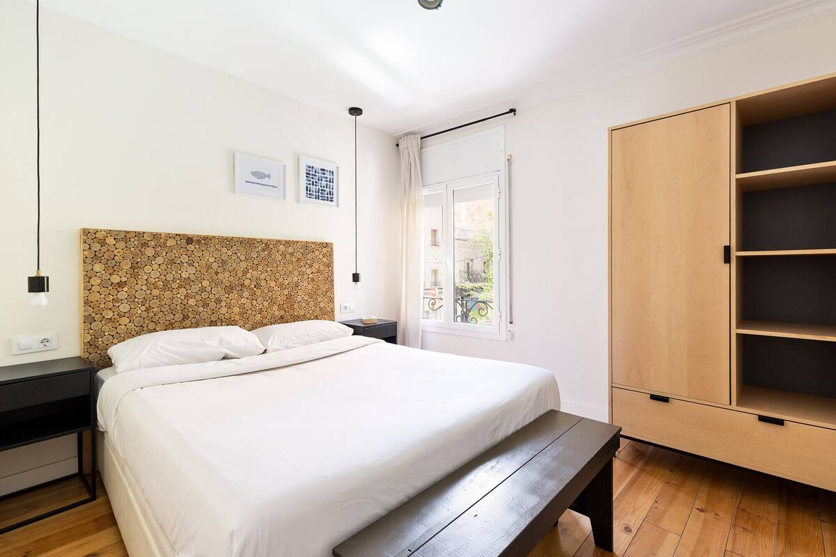 A bedroom with a bed, a wooden headboard, a nightstand, a bedside table, a wardrobe, and a window with a view of the outside.