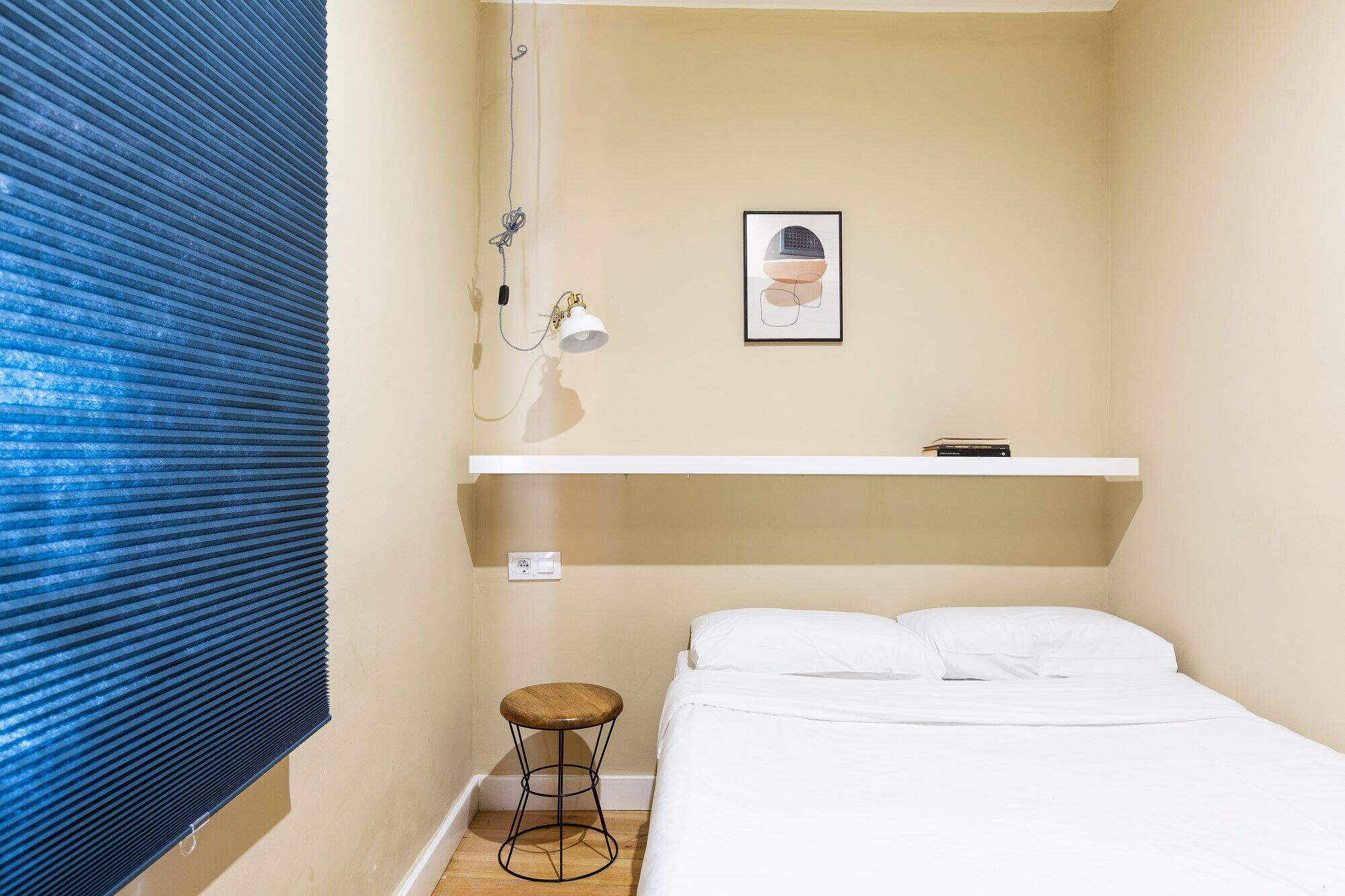 Un dormitorio pequeño y ordenado con una cama individual, un taburete, una estantería con libros y una lámpara fijada a la pared.
