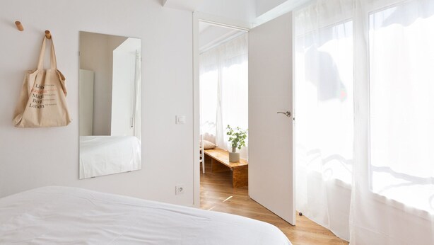 Une chambre à coucher lumineuse et épurée, avec un grand lit, un sac suspendu, un miroir et une vue sur un balcon, accessible par une porte ouverte.