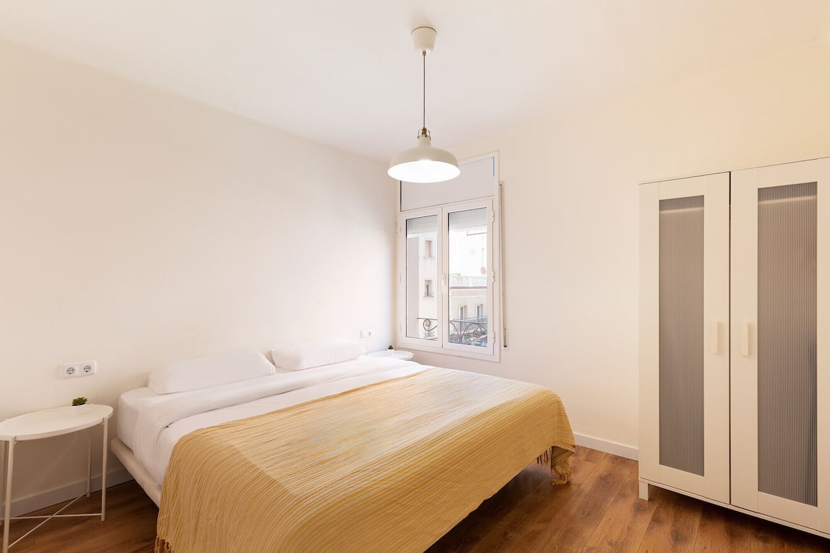A bedroom with a bed, bedside table, wardrobe, and a window with a view of buildings.
