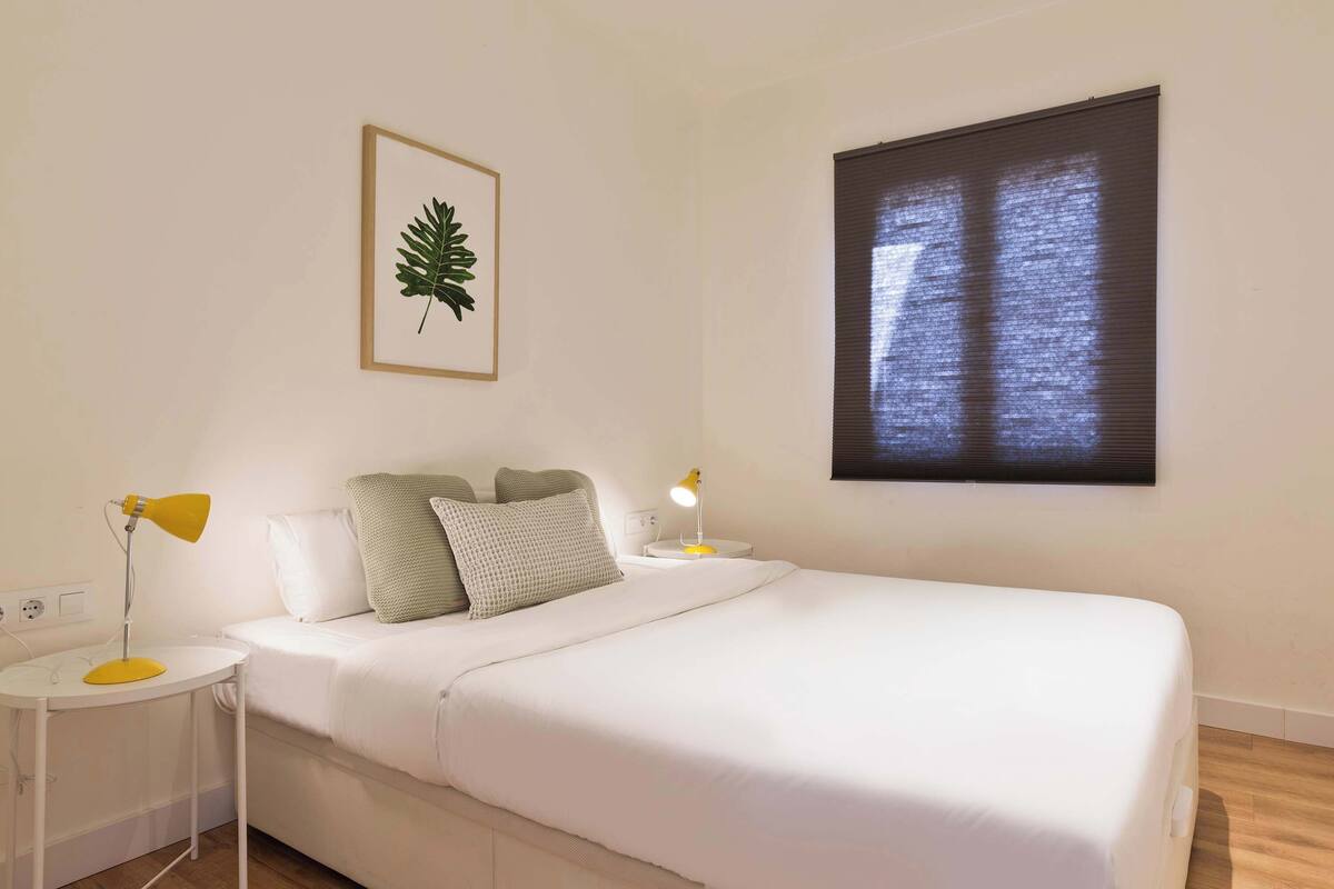 Un dormitorio con una cama, dos mesitas de noche, una obra de arte con una hoja enmarco, una ventana con persianas y un enchufe.