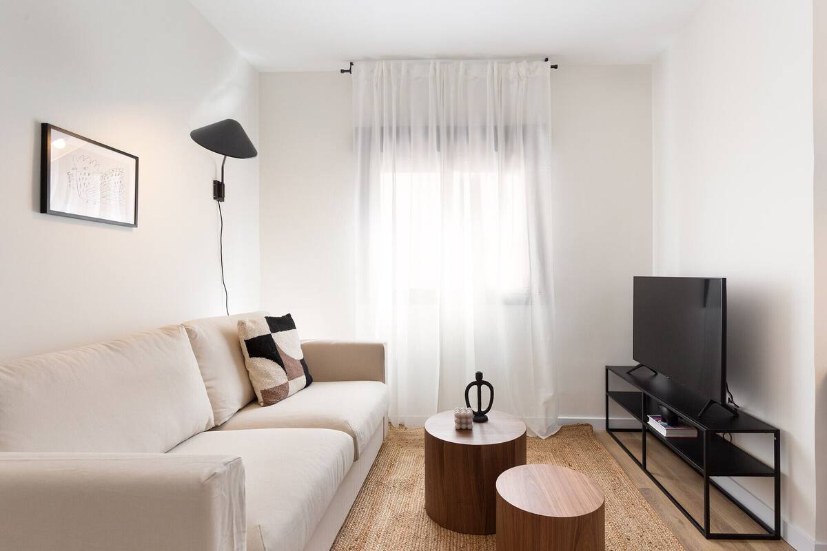 Un salón minimalista con un sofá, una mesa de centro, un mueble para TV y un ventanal con cortinas translúcidas.