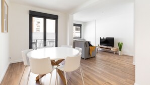 Un salon moderne avec une table à manger ronde, des chaises blanches, un canapé gris, un téléviseur à écran plat et une plante en pot.