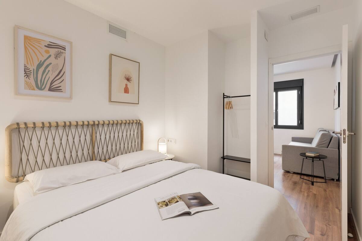 Un dormitorio moderno con una cama grande, una mesita de noche y una lámpara de lectura. Se ve una puerta que da a un área de estar con un sofá y una pequeña mesa.