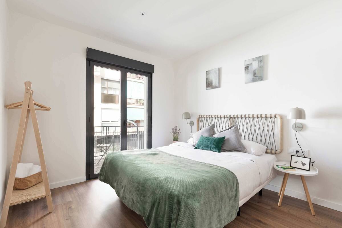 Un dormitorio con una cama, un perchero de madera, una ventana y una mesita de noche con una lámpara y un marco de fotos.