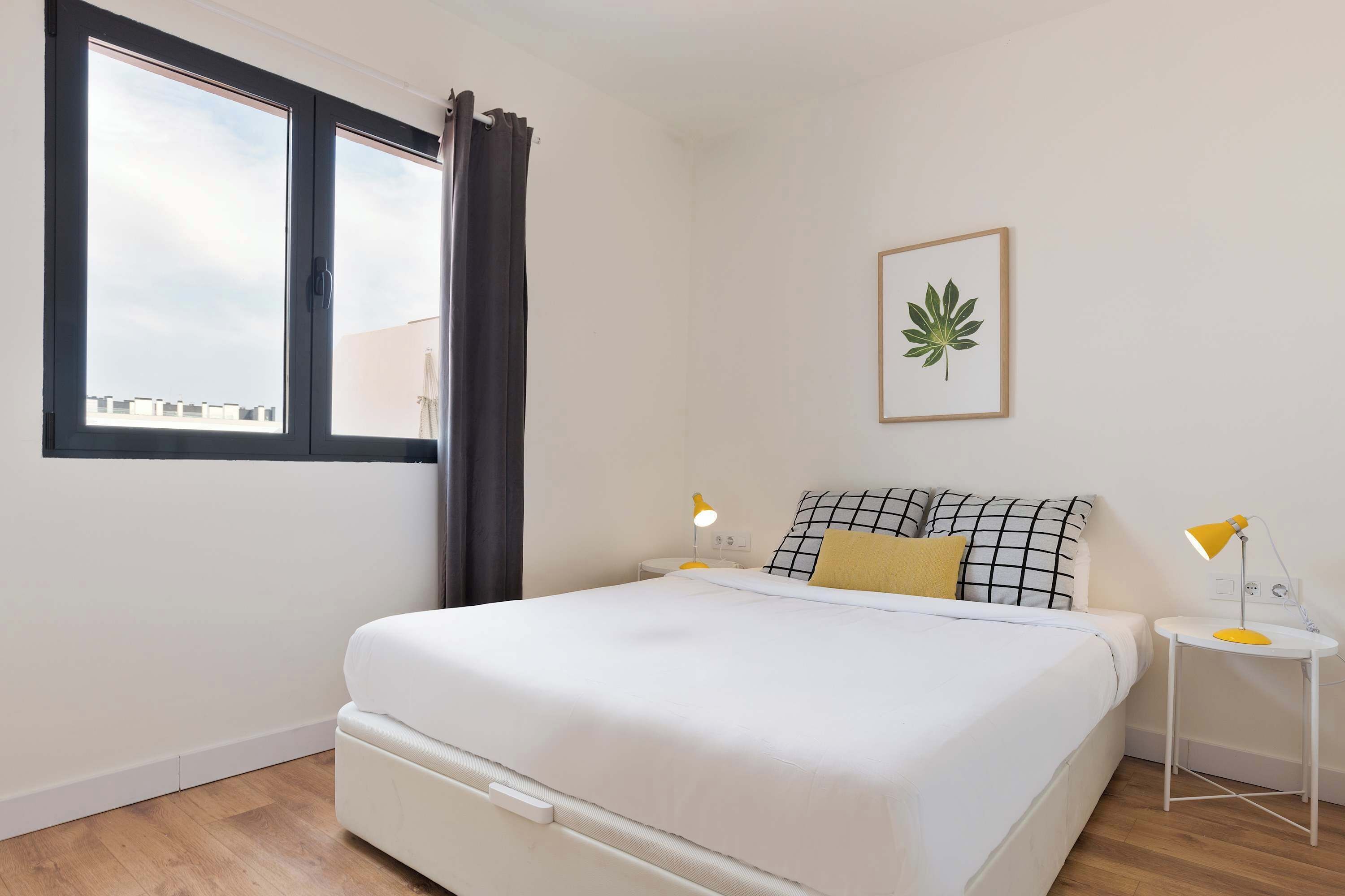 Un dormitorio con cama, mesita de noche, planta en la pared y ventana con cortinas.