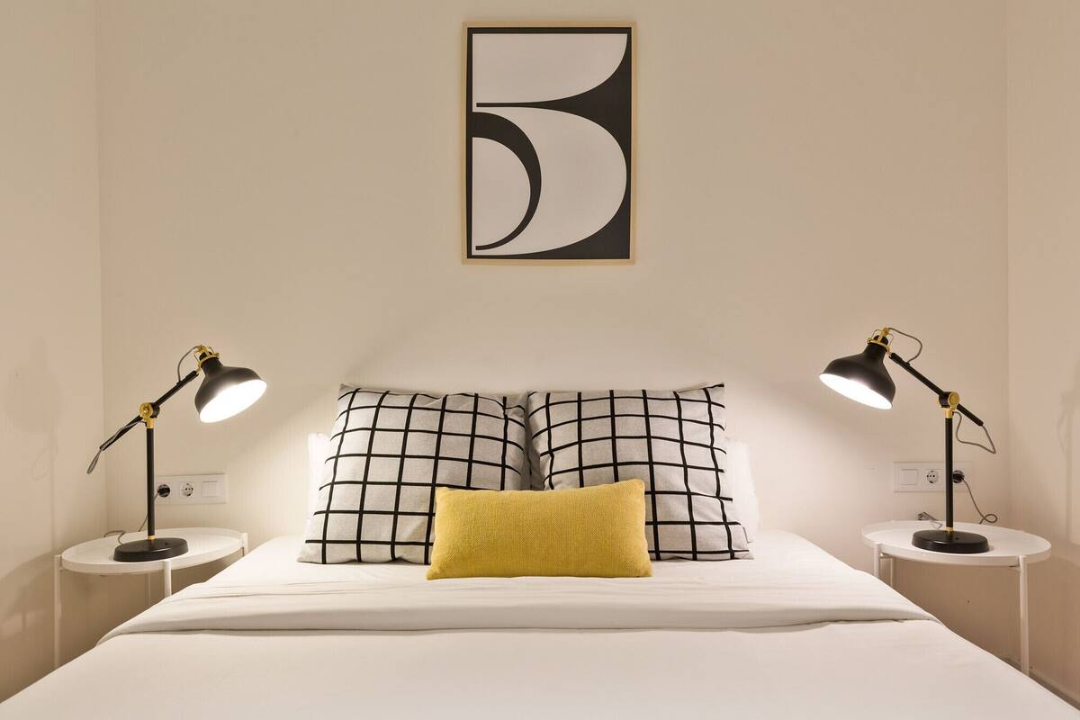 Una cama bien hecha con dos mesitas de noche, cada una con una lámpara, una gran obra de arte abstracta sobre la cama y un solo cojín amarillo.