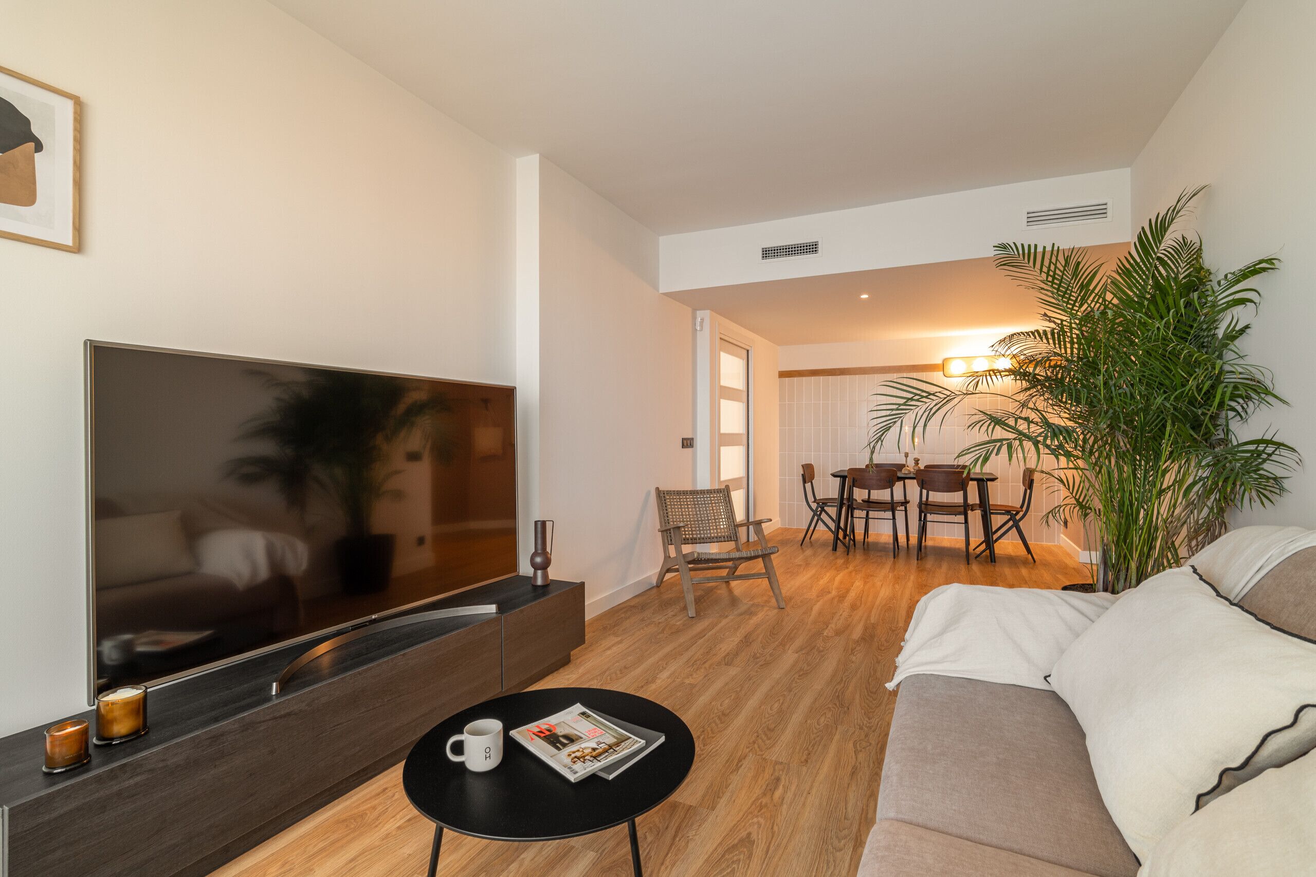 Una sala de estar moderna con un televisor de pantalla plana grande, una mesa de centro con una revista, un sofá y una zona de comedor con sillas y una mesa.