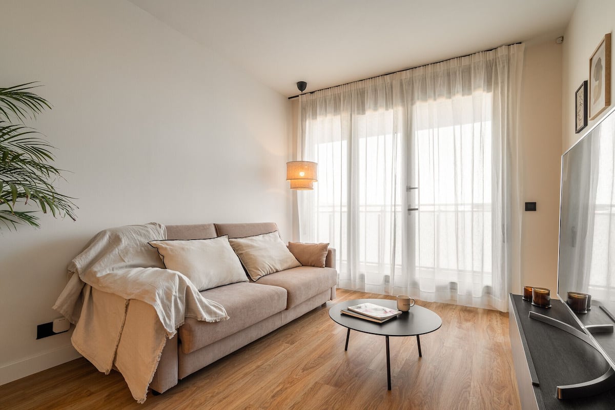 Un salon avec un canapé beige, une table basse ronde et une grande fenêtre ornée de rideaux légers.