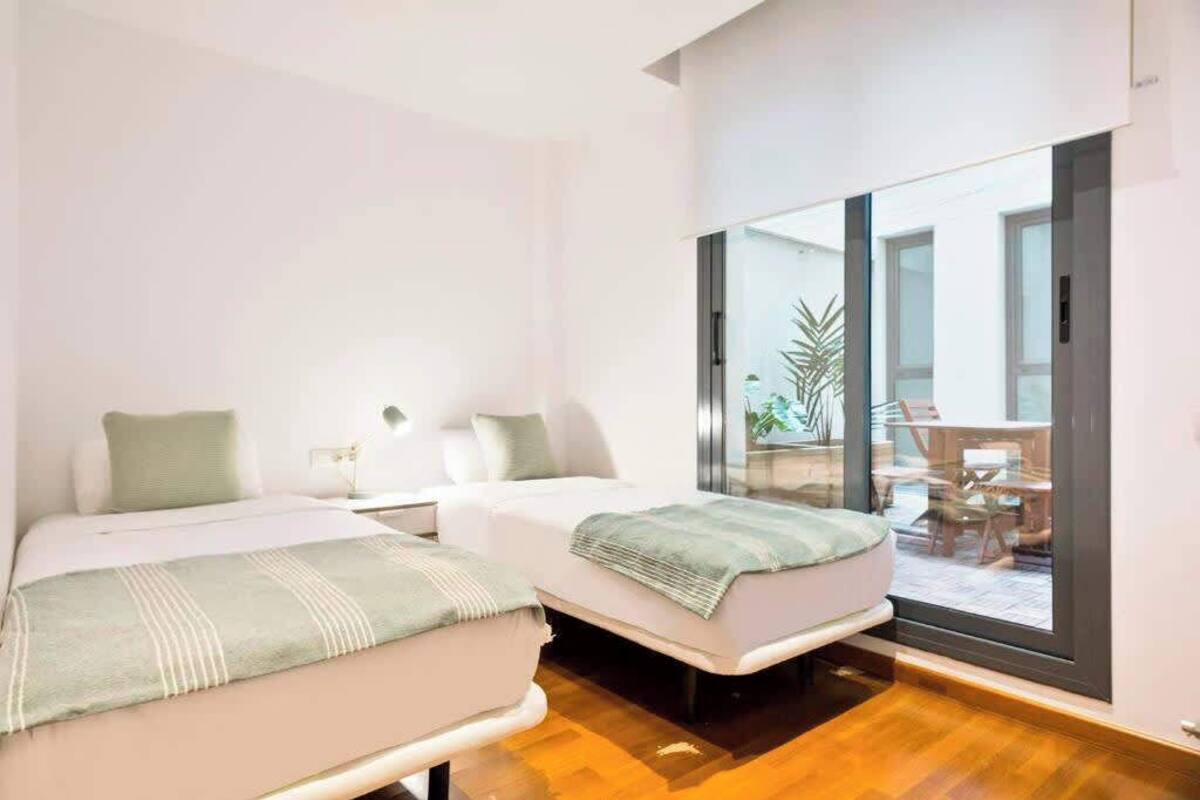 A modern hotel room with two beds, a large sliding glass door leading to an outdoor area, and a small table with a chair.