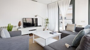 Una sala de estar moderna con un televisor de pantalla plana, muebles blancos y un amplio ventanal con cortinas.