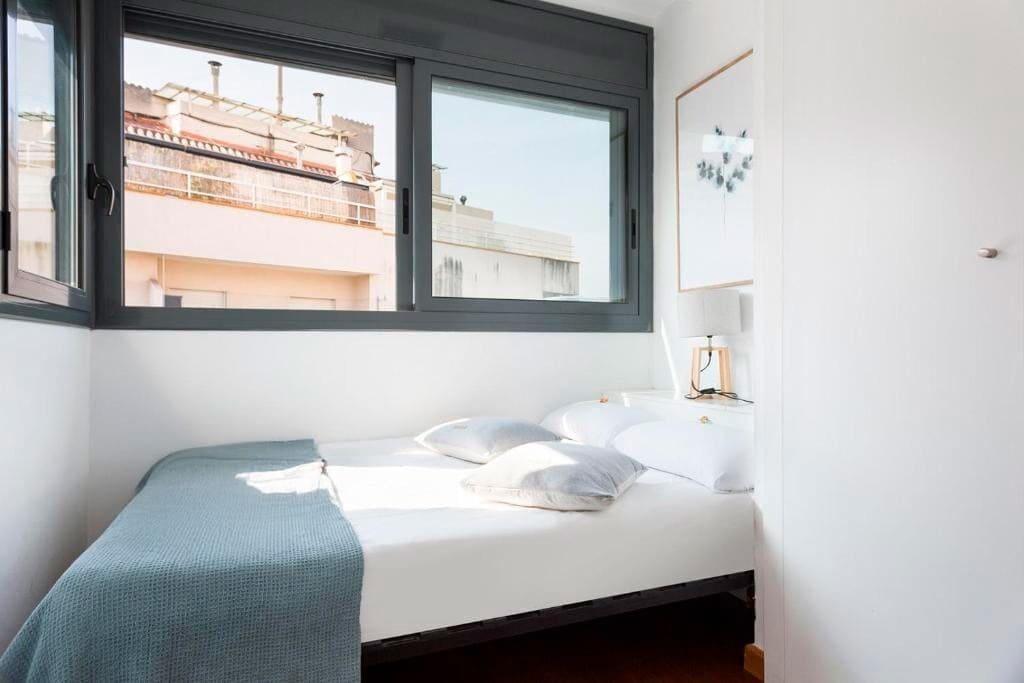A bedroom with a bed, pillows, a bedside lamp, and a window with a view of buildings.