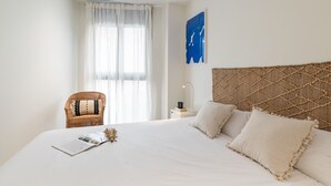 Un dormitorio ordenado con una cama grande, una silla de mimbre, una mesita de noche y una obra de arte colgada en la pared.