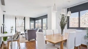 Una habitación moderna y bien iluminada con una mesa de comedor de madera, sillas blancas y un sofá gris. Amplias ventanas ofrecen vistas del paisaje urbano.