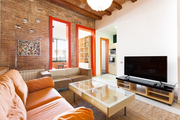 A living room with a brick wall, orange-framed windows, a sofa, a coffee table, and a TV.