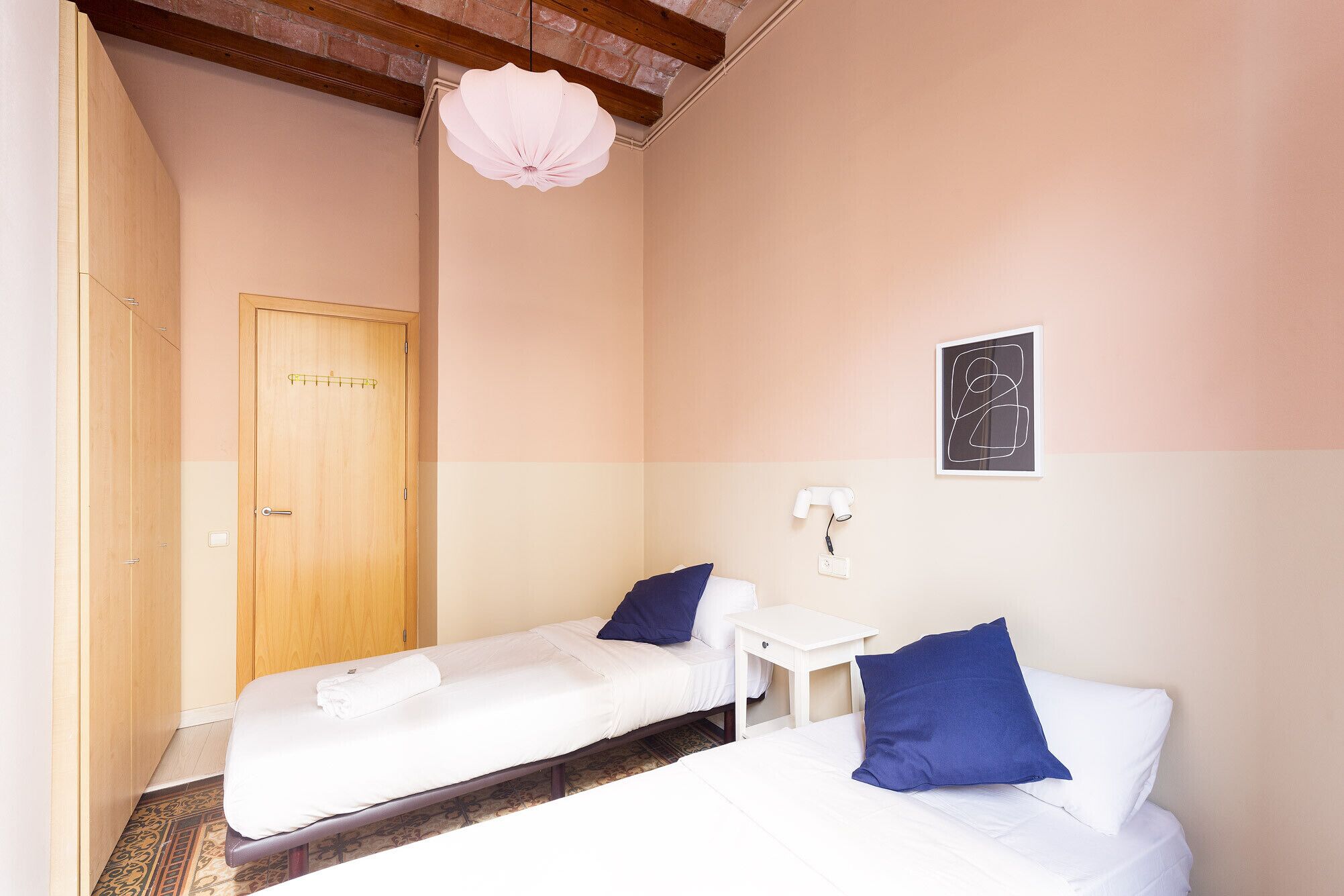 Une chambre avec deux lits simples, une porte en bois, une table de chevet blanche, une lampe fixée au mur et une œuvre d’art abstraite encadrée.
