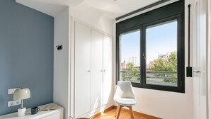Una habitación con un armario blanco, una silla blanca, un ventanal con marco negro y vistas a edificios y árboles.