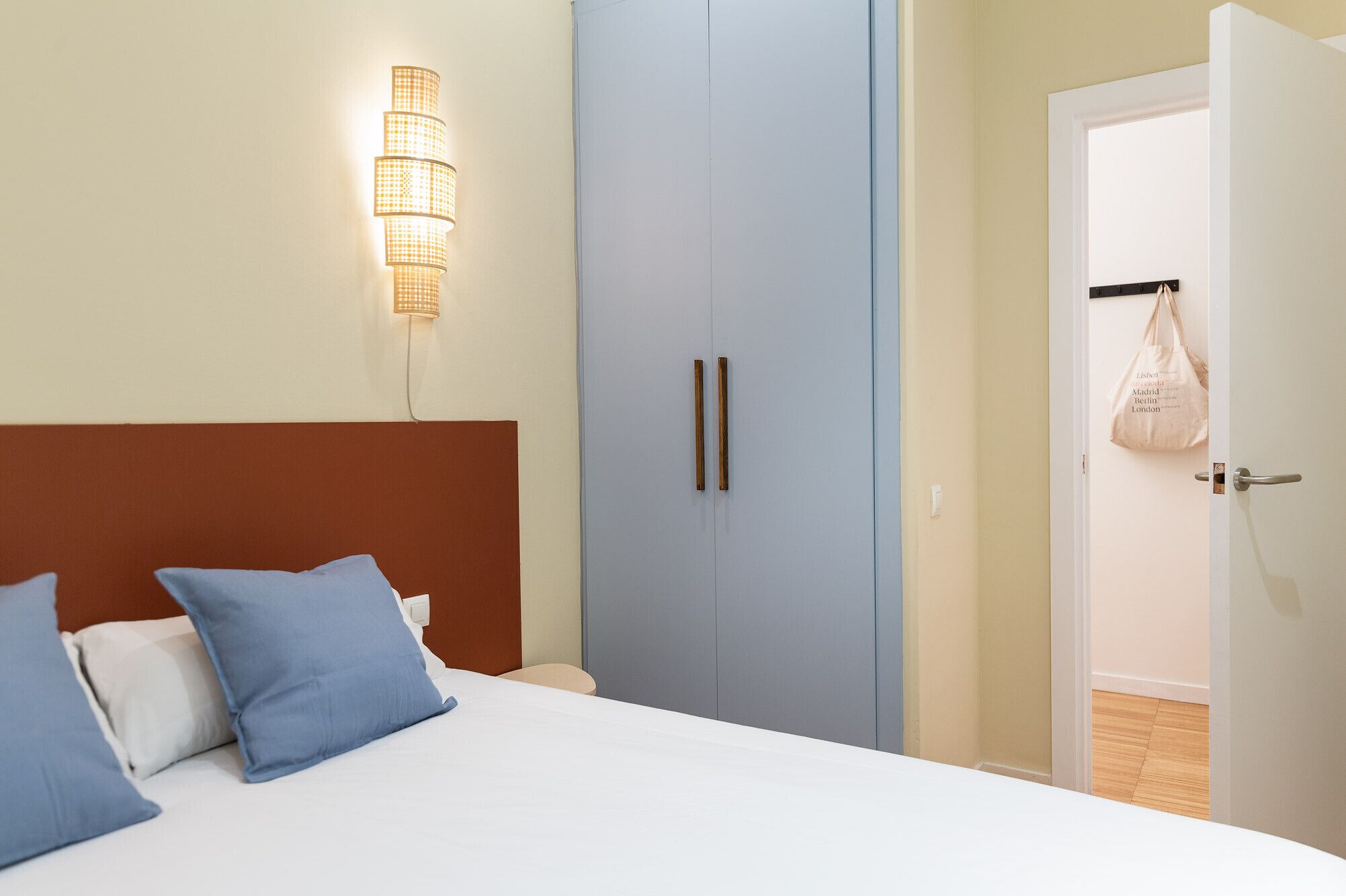 A bedroom with a bed, two pillows, a wall-mounted light fixture, a wardrobe, and a door with a bag hanging on it.