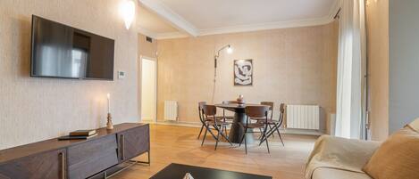 Een moderne woonkamer met een flatscreen televisie, een eethoek met een ronde tafel en stoelen, en een bank.