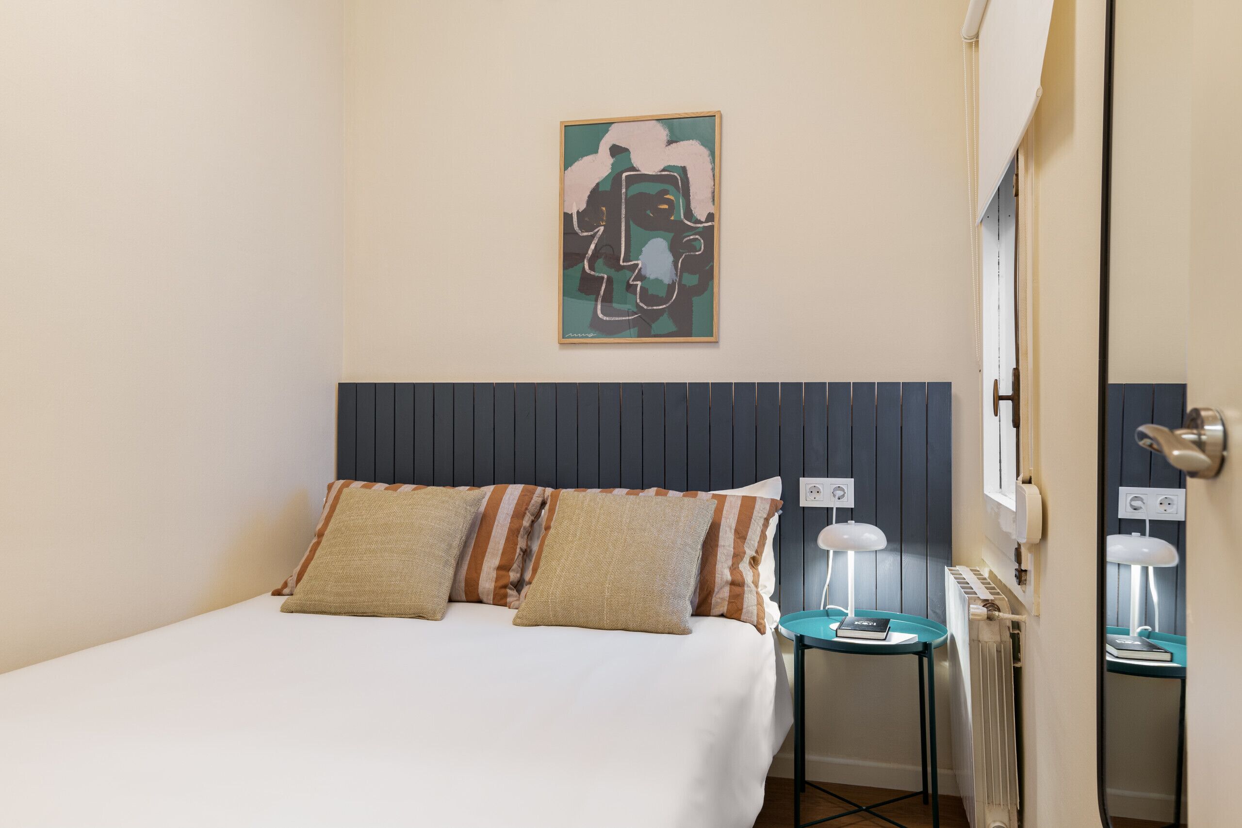 Une chambre d’hôtel avec un lit, deux oreillers, une table de chevet avec une lampe, un radiateur et une œuvre d’art encadrée au mur.