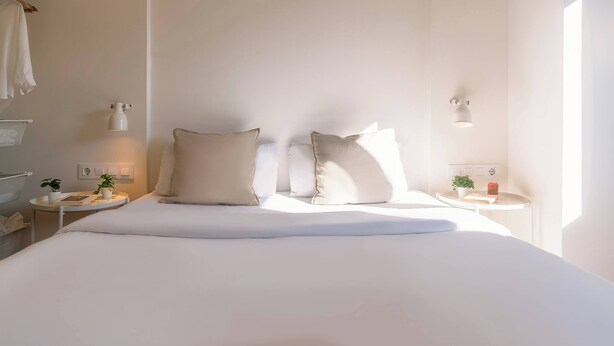 Un dormitorio minimalista con una cama grande, dos mesitas de noche con lámparas y una mesita más pequeña con una planta, y vistas al baño.
