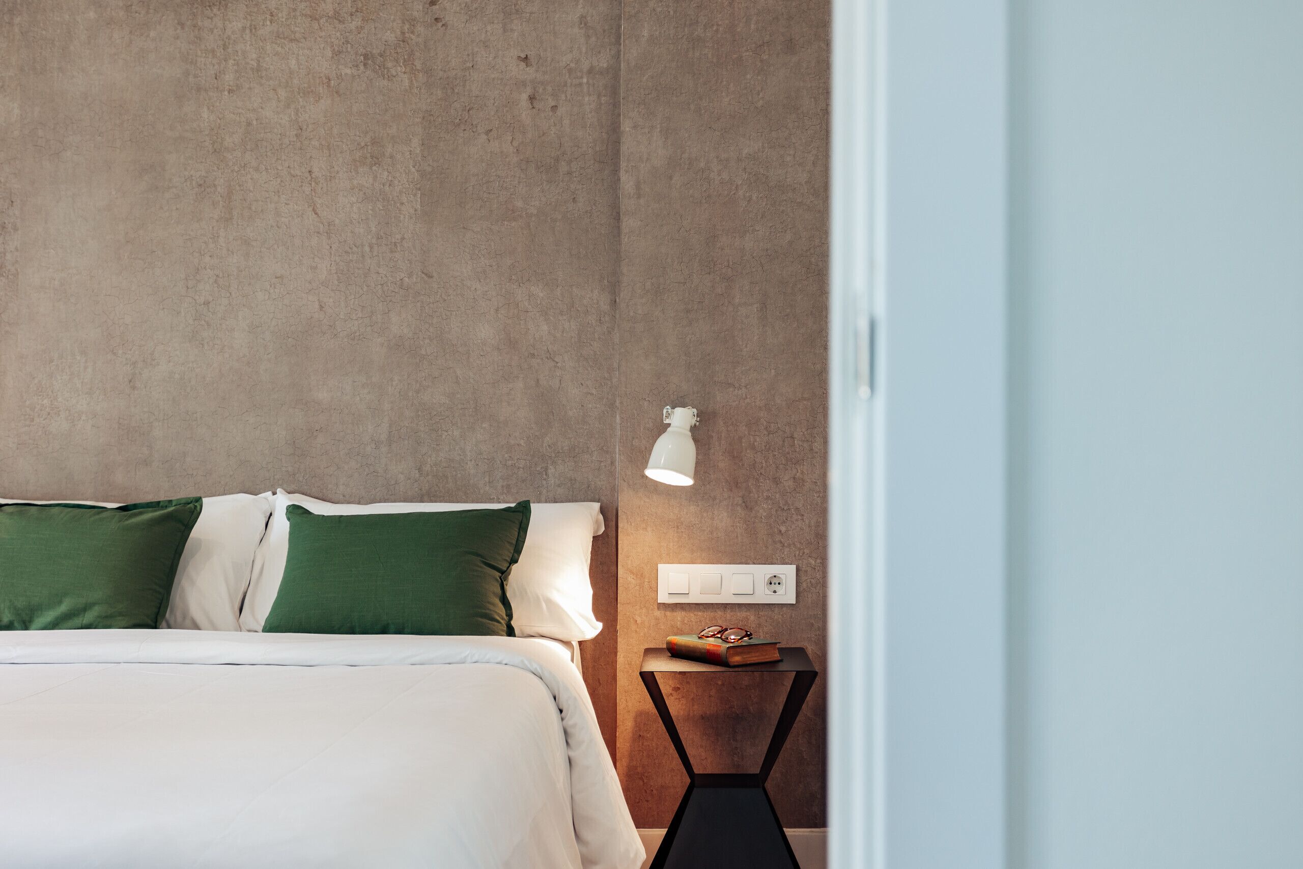 Un dormitorio con una cama, almohadas verdes y blancas, una mesita de noche con un libro y gafas, y una lámpara de pared.