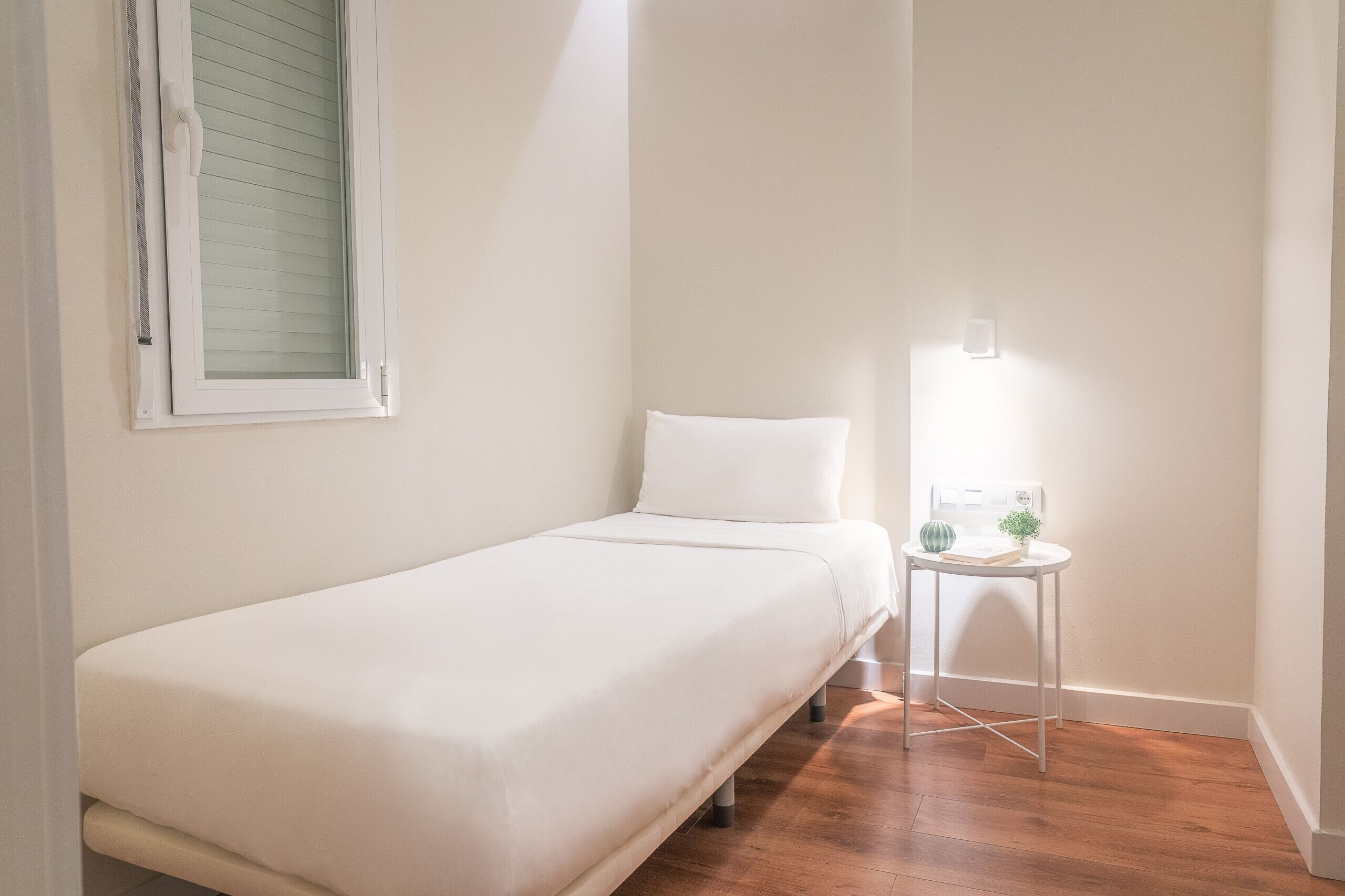 Un petit lit, bien fait, dans une pièce au plancher de bois, avec une petite table et une lampe, et une fenêtre munie de stores.