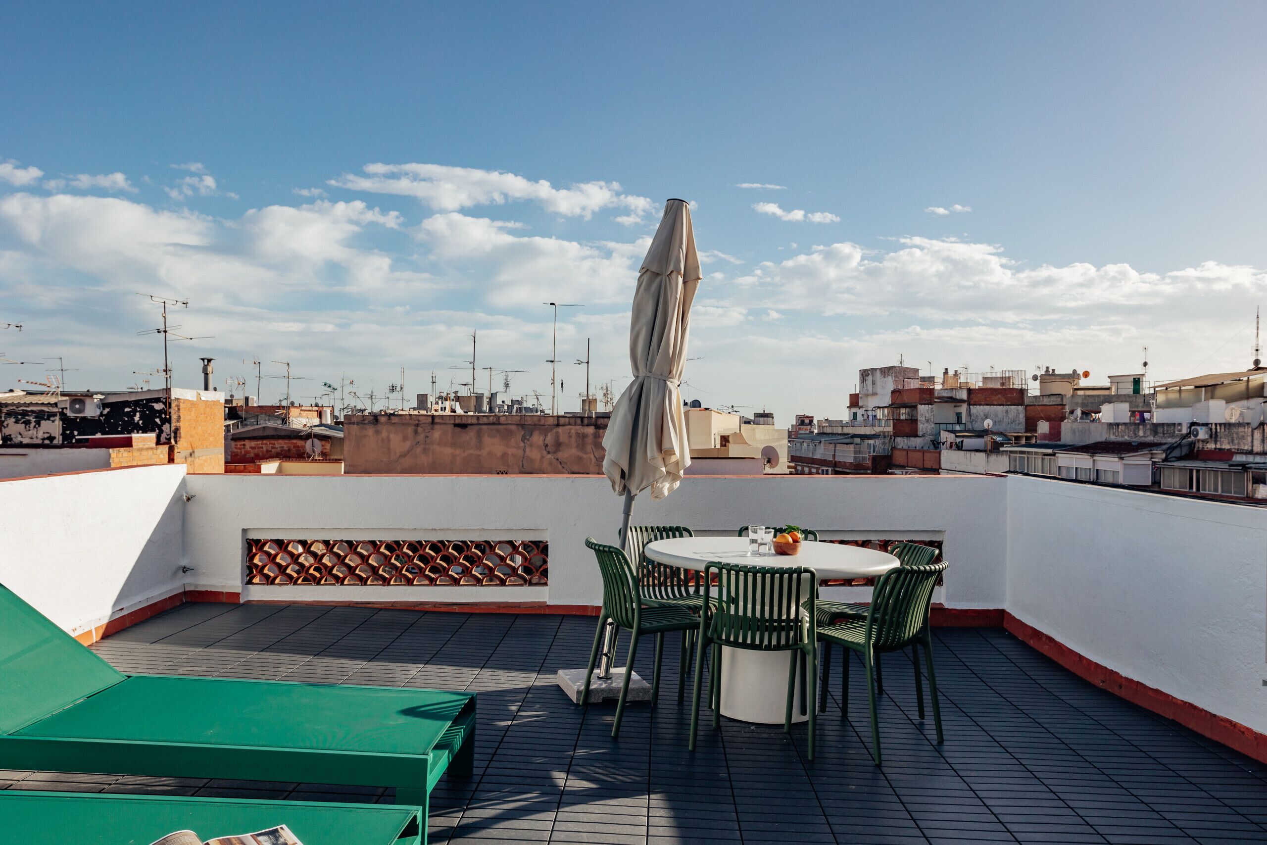 Una terraza en la azotea con una mesa redonda y sillas, una zona de estar verde, y vistas del paisaje urbano.