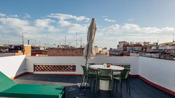Una terraza en la azotea con una mesa redonda y sillas, una zona de estar verde, y vistas del paisaje urbano.