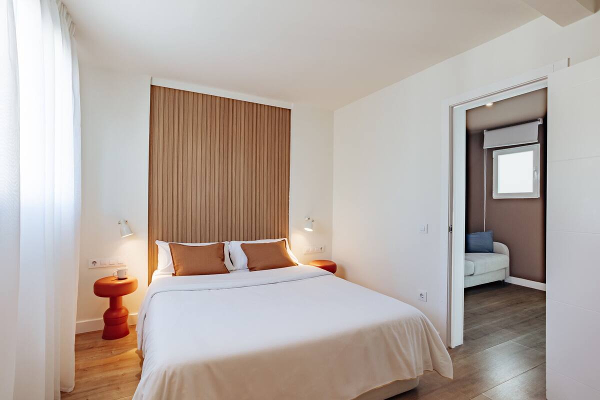 Un dormitorio con una cama grande, cabecera de madera, lámparas de noche y vistas a una sala de estar a través de una puerta abierta.