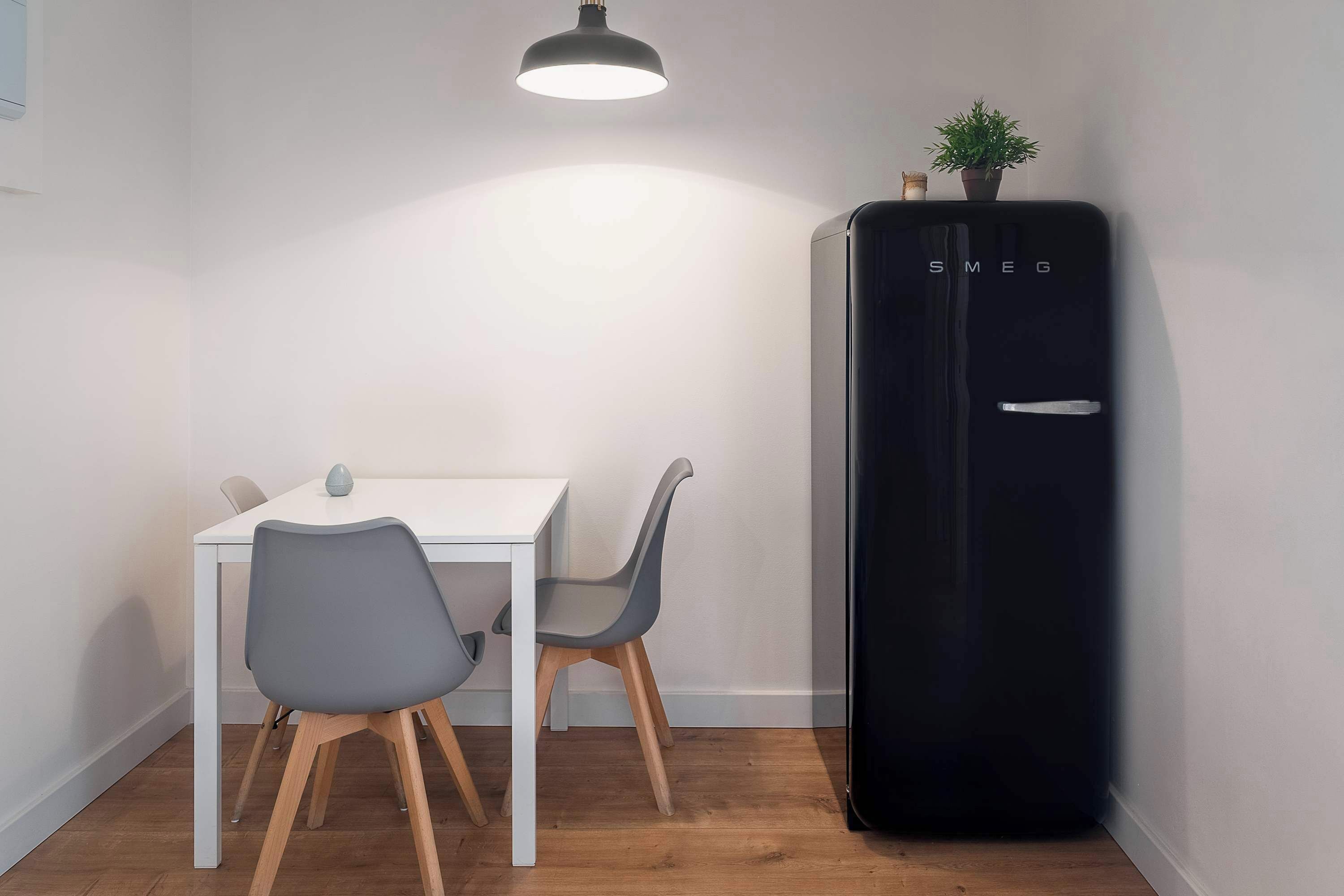 A compact room with a white table and two grey chairs, a black refrigerator, and a hanging light fixture.