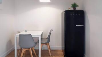 A compact room with a white table and two grey chairs, a black refrigerator, and a hanging light fixture.